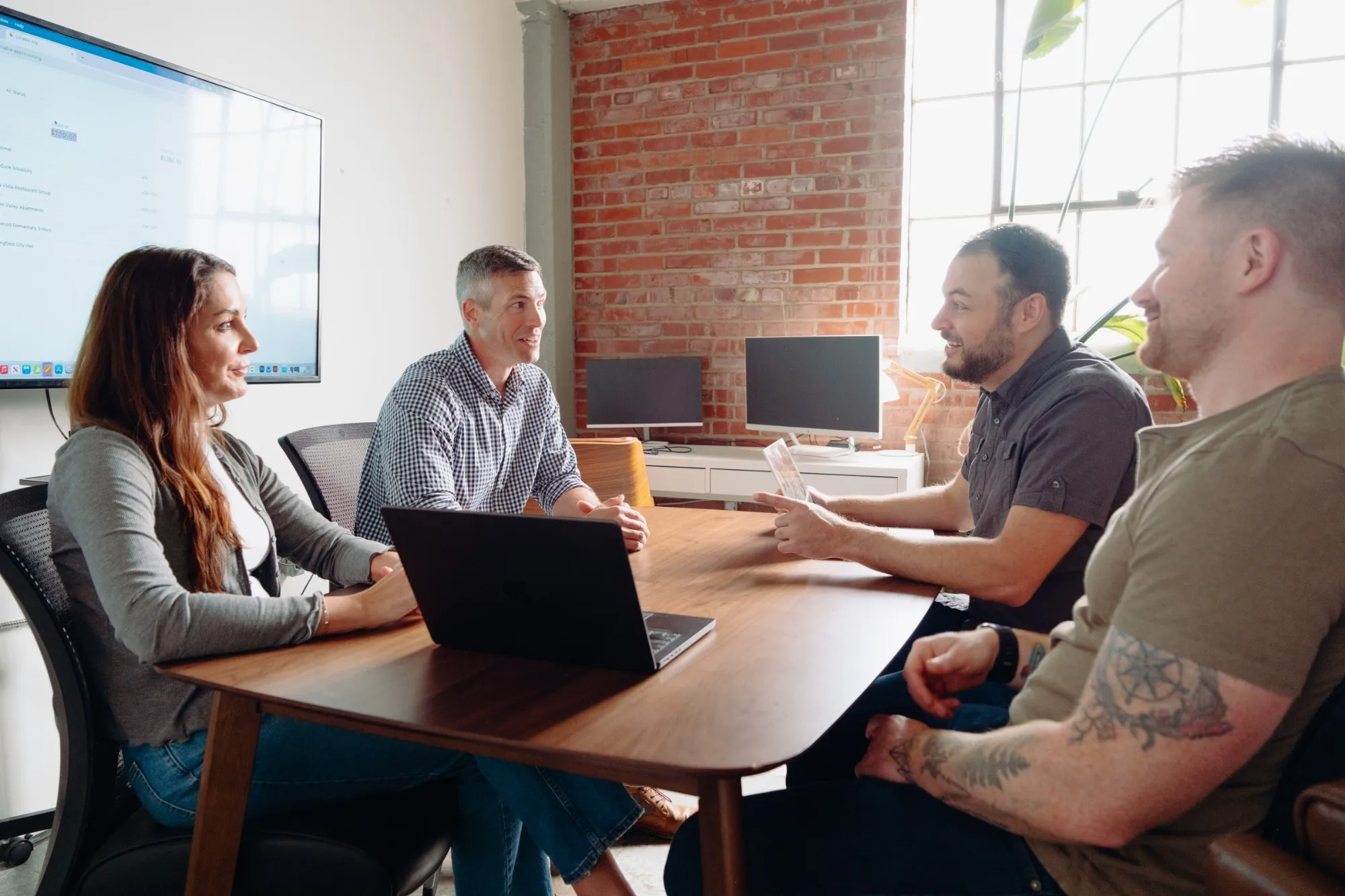 Four people in a working session around a conference table, with an app up on the wall screen